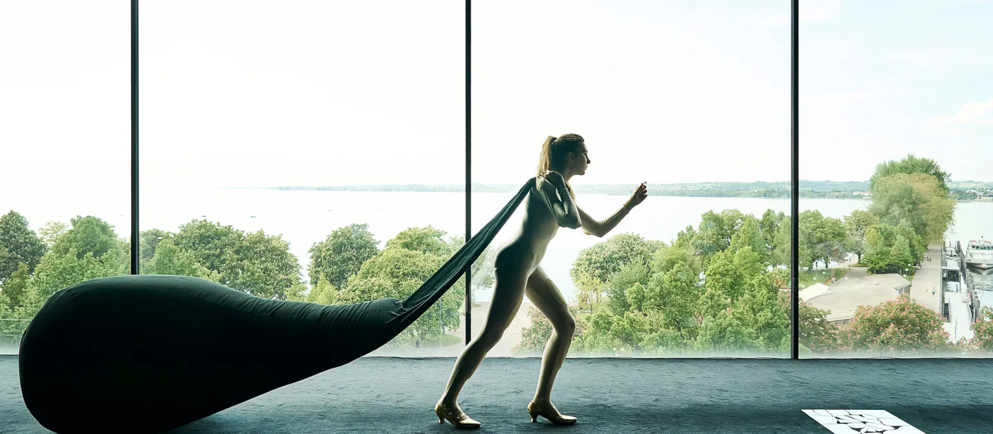 Szene aus "Ihr seid bereits eingeschifft", 2021, diverse Spielorte, Frau zieht Sack hinter sich her, vor Panoramafenster mit Blick auf Bäume und Wasser