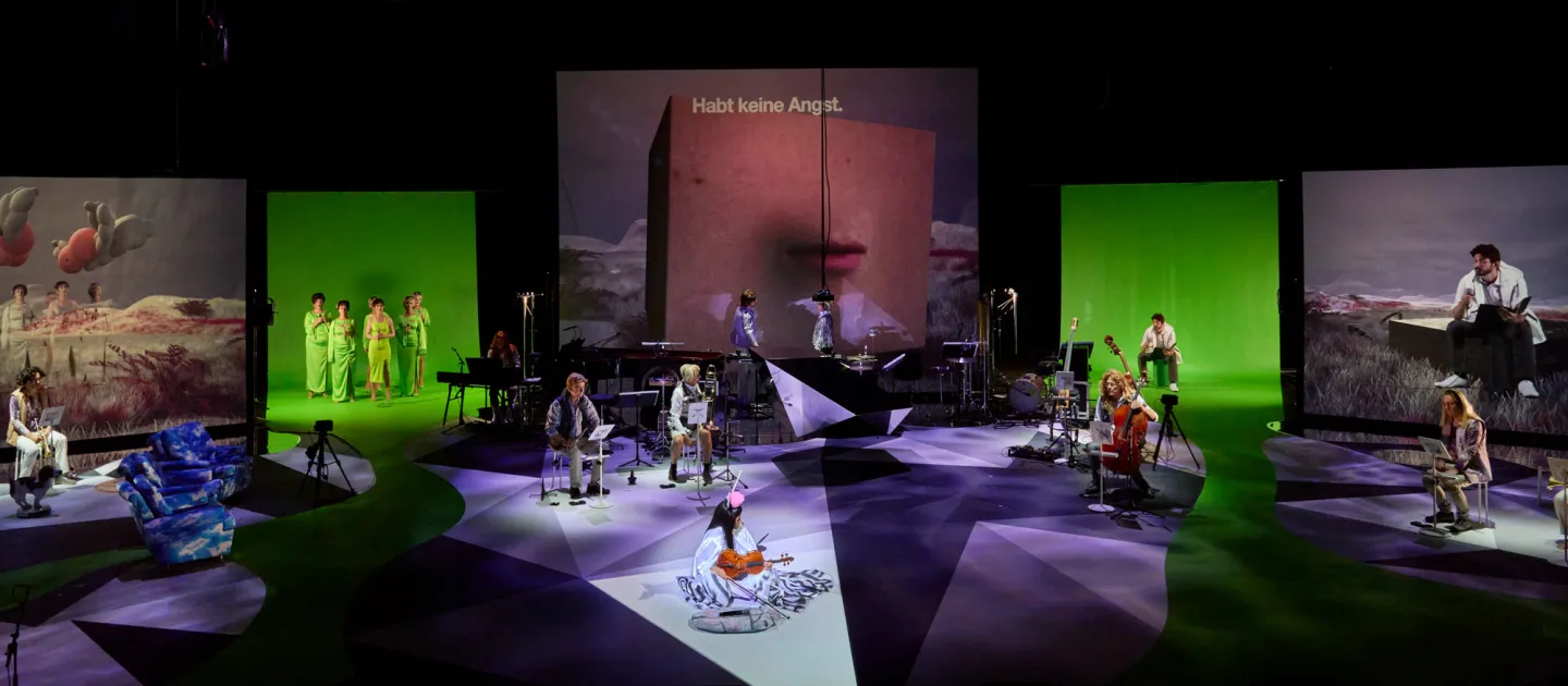 Szene aus "Melencolia", 2022 im Festspielhaus, mehrere kostümierte Darstellende und Musiker:innen auf der Bühne verteilt, große Bildschirme im Hintergrund, mittig die Aufschrift "Habt keine Angst"