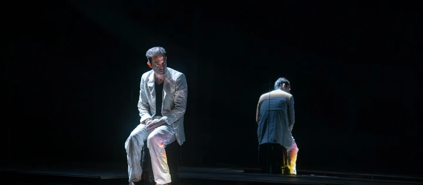 Szene aus "Der Menschenfeind", 2024 im Festspielhaus, ein sitzender Darstellender in hellem Anzug, Spiegelung der Person im Hintergrund