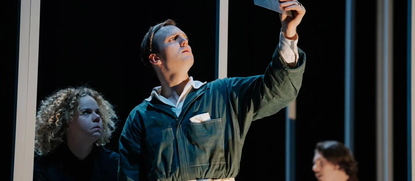 Szene aus "Richard II.", 2021 im Festspielhaus, drei Darstellende, zentral ein Darstellender in dunkelgrünem Overall mit hochgehaltenem Dokument in der Hand