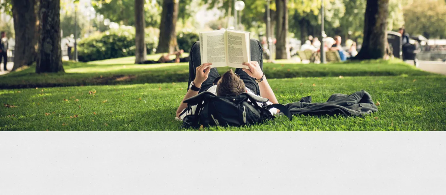 Person liegt auf dem Rücken im Park und liest ein Buch, umgeben von Bäumen und Gras