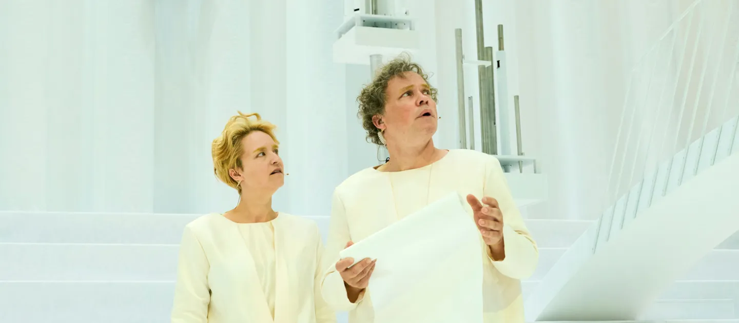 Szene aus "Wind", 2021 im Festspielhaus, zwei Darstellende in weißem Kostüm, Blick nach links richtend, eine Person hält ein großes Blatt Papier, heller Hintergrund mit Treppen