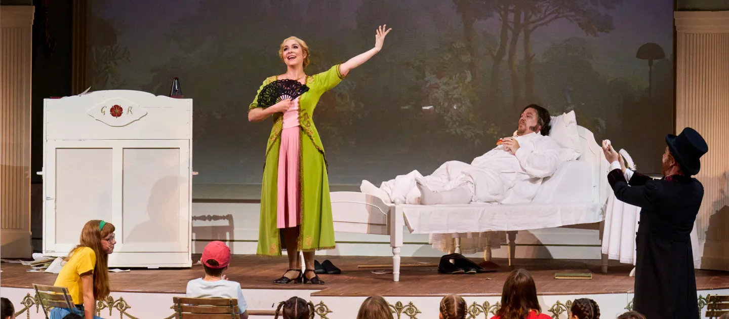 Szene aus "Die Zeitreisemaschine", 2022 im Festspielhaus, Darstellende in grün-rosafarbenem Kostüm mit schwarzem Fächer in der rechten Hand, ein Mann liegt im Bett, Kinder im Publikum