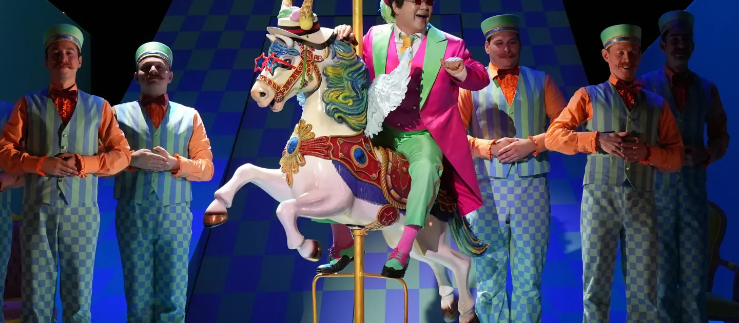 Ein Mann in einem bunten Anzug mit einem Zylinder reitet auf einem weißen Karussellpferd. Um ihn herum stehen mehrere Männer in gestreiften Westen und Krawatten.