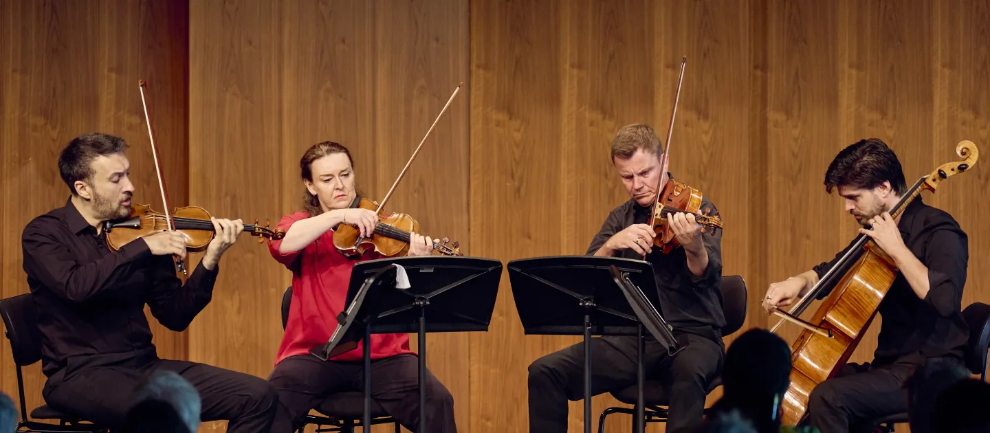 Vier Musiker in schwarzer Kleidung spielen ein Streichquartett auf der Bühne. Zwei Violinisten, ein Bratschist und ein Cellist stehen in einer Reihe hinter Notenständern.