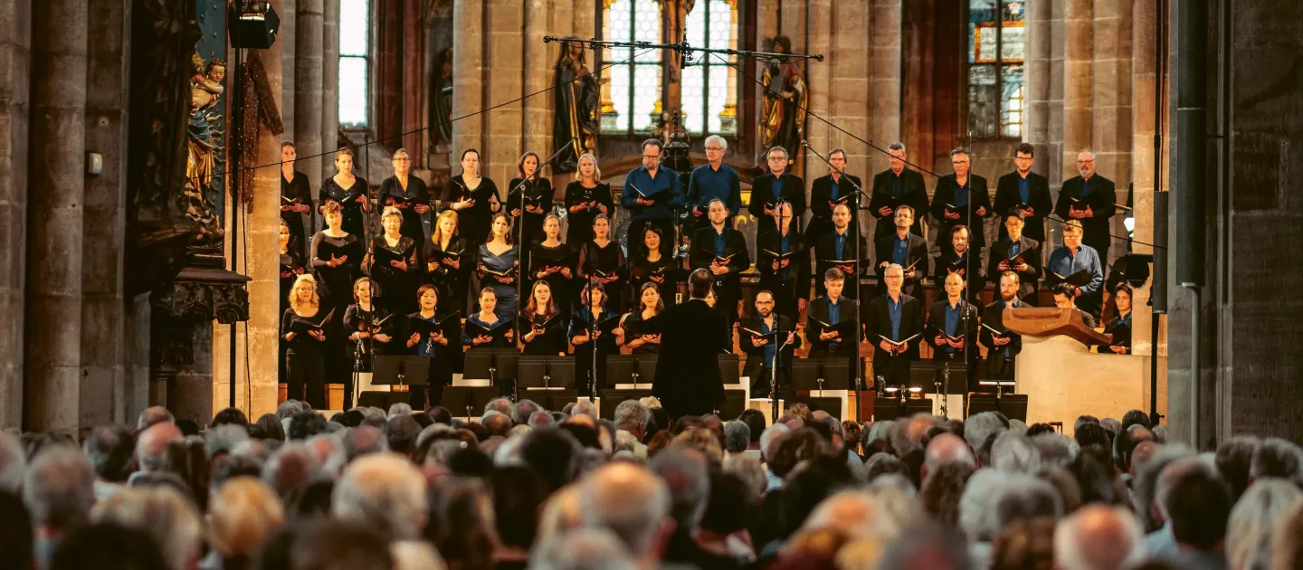 Großer Chor mit mehreren Sängerinnen und Sängern in dunkler Kleidung steht auf Stufen in einer Kirche vor Publikum, Dirigent mit erhobenen Armen.