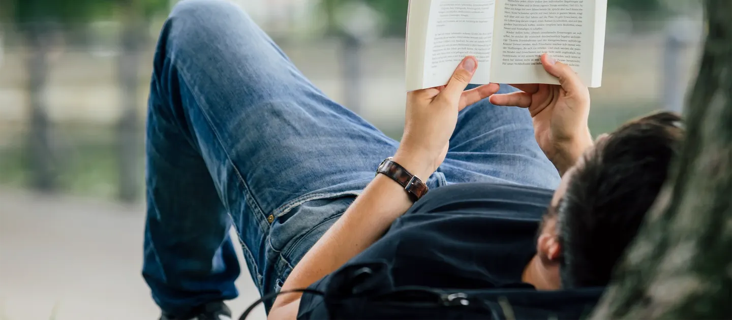 Person liegt entspannt auf dem Rücken und liest ein aufgeschlagenes Buch im Freien