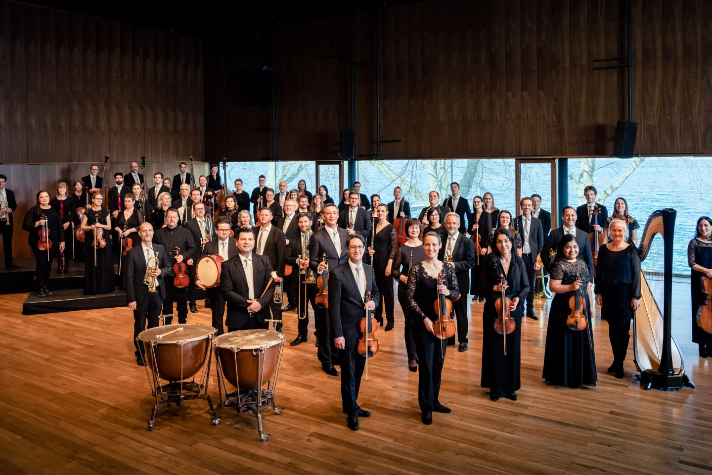 Symphonieorchester Vorarlberg mit Musikerinnen und Musikern in formeller Kleidung, stehend mit ihren Instrumenten in einem großen Konzertsaal mit Holzwänden und großen Fenstern