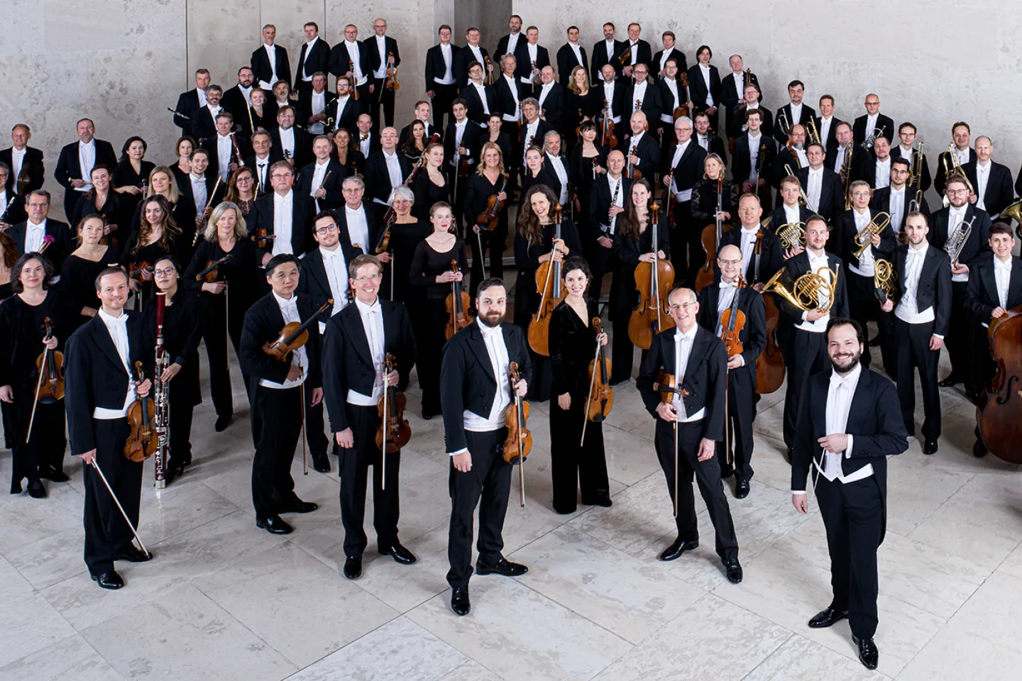 Mitglieder der Wiener Symphoniker in formeller Konzertkleidung mit Instrumenten, auf einer Treppe in einem modernen Saal gruppiert
