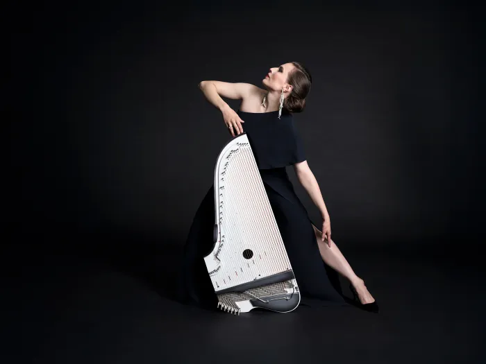 Ida Elina in schwarzem Kleid posiert elegant mit einer weißen Kantele vor schwarzem Hintergrund