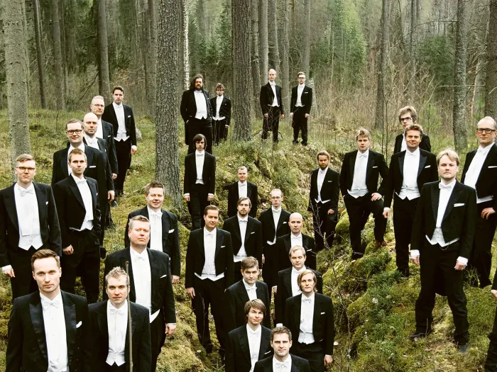 Männerchor YL Male Choir in schwarzem Smoking und weißem Hemd steht verteilt im Wald zwischen Bäumen und Moos