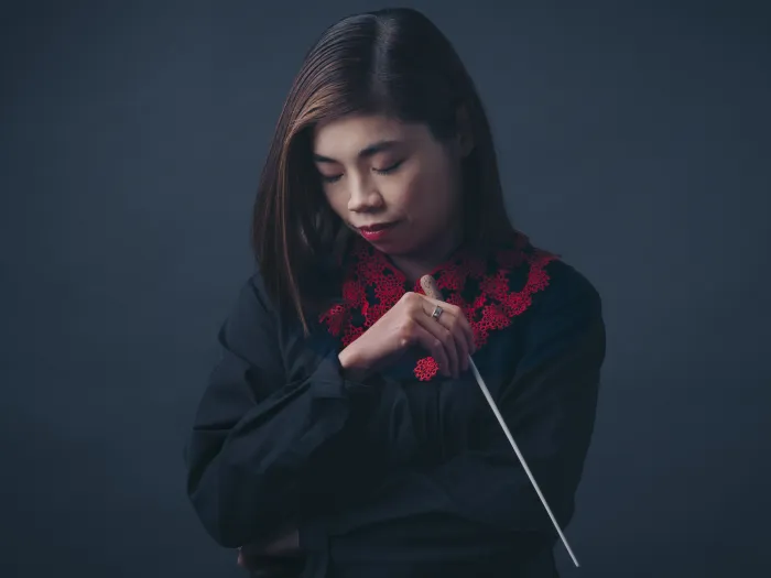 Dirigentin Elim Chan konzentriert mit geschlossenen Augen, hält Taktstock vor dunklem Hintergrund, trägt schwarzes Oberteil mit rotem Muster