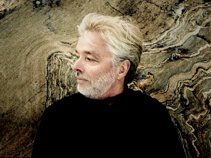 Jukka Pekka Saraste mit grauem Haar und Bart, vor einer strukturierten Felsoberfläche, trägt schwarzen Pullover, Blick nach links