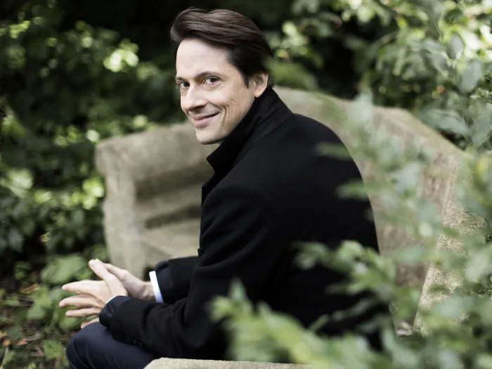 Dirigent Leo McFall sitzt auf einer Steinbank im Grünen, trägt einen schwarzen Mantel und lächelt leicht zur Seite