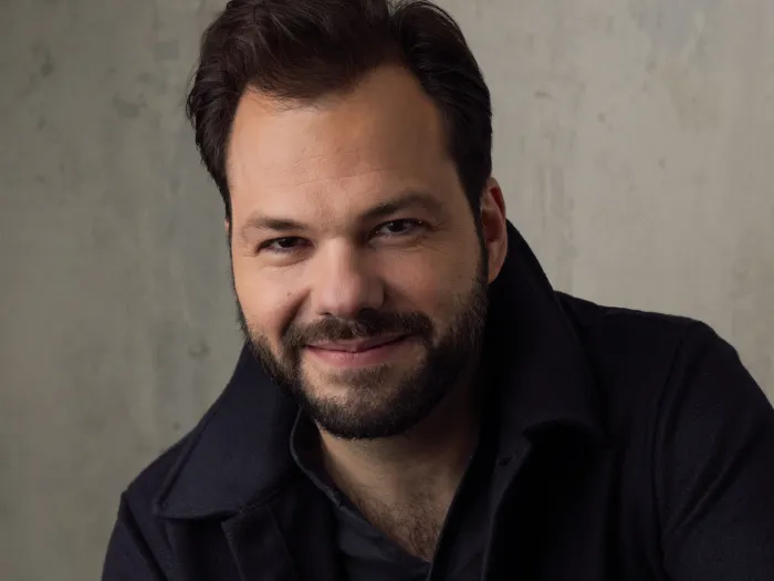 Porträt von Dirigent Petr Popelka mit kurzem dunklem Haar und Bart, lächelnd, vor hellem Hintergrund