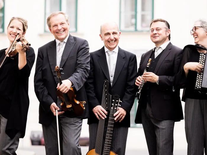 Fünf Musiker in schwarzer Kleidung stehen lachend mit ihren Instrumenten auf einer Straße, darunter Geige, Gitarre, Klarinette und Akkordeon.