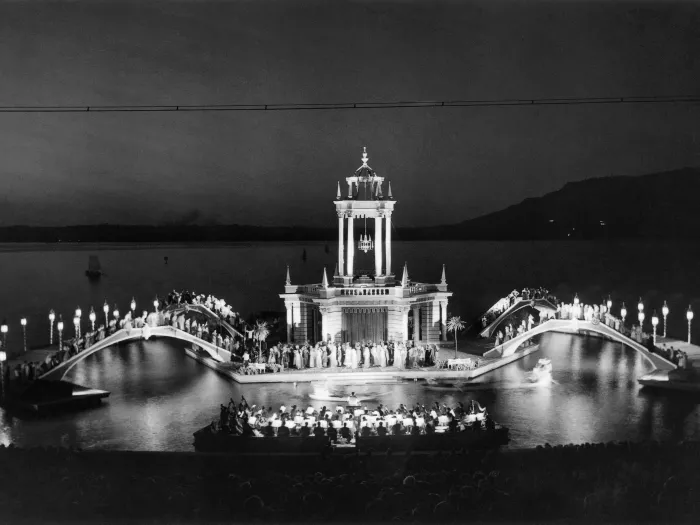 Szene aus Boccaccio, 1953 auf der Seebühne, viele Menschen, im Mittelpunkt befindet sich eine Insel, auf der sich ein Gebäude mit Turm befindet, links und rechts schließt sich je eine gebogene Brücke an, im Vordergrund gibt es ein Boot 