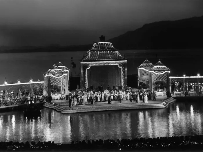 Szene aus Fledermaus, 1954 auf der Seebühne, viele Menschen, im Mittelpunkt befindet sich ein großer Pavillon, links und rechts stehen je zwei weitere, kleinere Pavillons, von denen je ein Steg abgeht