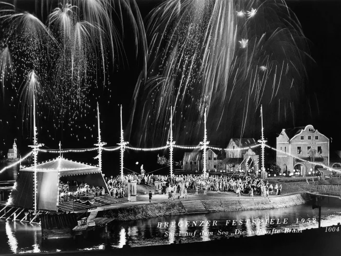Szene aus "Die verkaufte Braut", 1958 auf der Seebühne, links befindet sich ein Festzelt mit einer Menschengruppe, rechts daneben liegt ein Festplatz, gefüllt mit Menschen, im rechten Hintergrund ist eine Häuserreihung zu sehen 