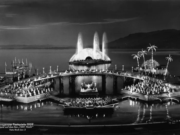 Szene aus "Tausendundeine Nacht", 1959 auf der Seebühne, im Vordergrund befindet sich mittig eine kleine Plattform mit einer kleinen Gruppe von Menschen, links und rechts daneben liegen je eine größere Plattform mit einer großen Menschenmenge, die durch einen höher gelegenen Steg miteinander verbunden sind, hinter dem Steg sind auf der linken Seite Schiffsmasten zu sehen, auf der rechten Seite ein Gebäude und Palmen, im Hintergrund ist eine weitere Plattform mit großem Wasserspiel zu sehen