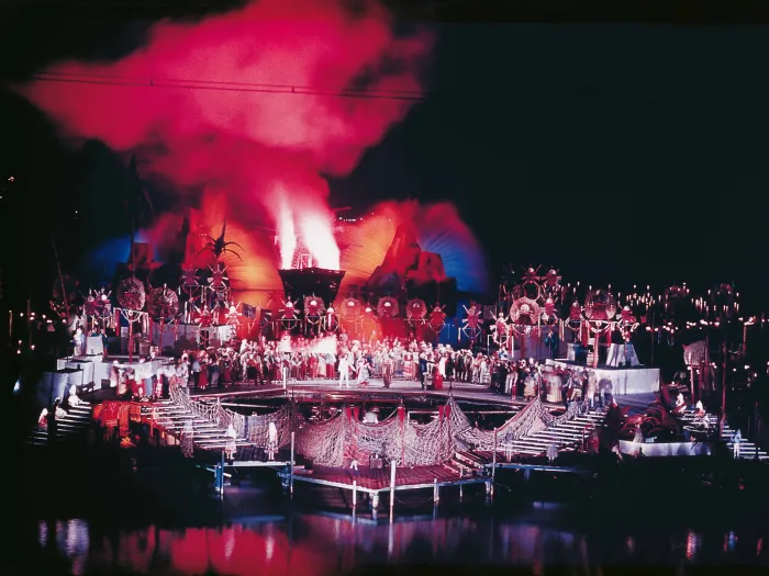Szene aus "Trauminsel", 1962 auf der Seebühne, viele Darstellende in Kostümen im Vordergrund, dahinter erhöhte Verzierungen, im Hintergrund ein Wasserspiel, das in Rot und Blau beleuchtet ist, roter Rauch steigt auf