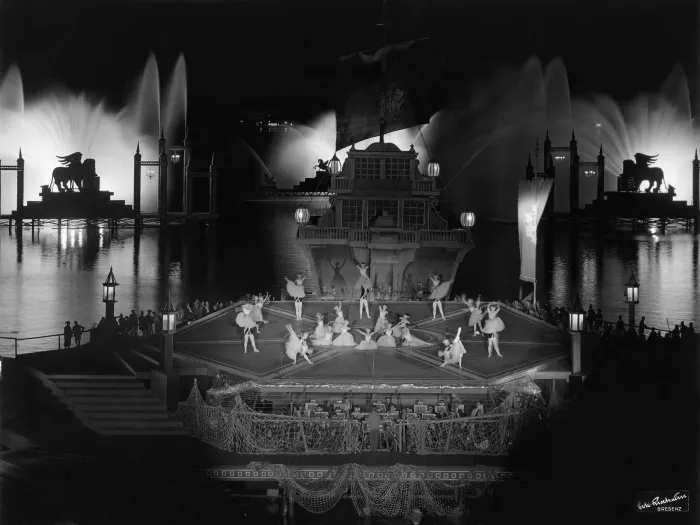 Szene aus "Eine Nacht in Venedig", 1965 auf der Seebühne, mittig befindet sich eine Plattform in Form eines Hexagon mit verschiedenen Balletttänzer:innen, an der Rückseite ist ein mehrstöckiges historisch-aussehendes Schiff angeschlossen, im Hintergrund ist links und rechts eine weitere Plattform mit je einer Silhouette eines geflügelten Löwen (Markuslöwe) und mittig eine Plattform mit einem geflügelten Pferd (Pegasus) zu sehen