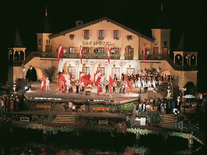 Szene aus "Hochzeit am Bodensee", 1969 auf der Seebühne, eine eckig gestaltete Plattform, auf der ein zweistöckiges Gebäude mit der Aufschrift "Seehotel" steht, zahlreiche Darstellende, teils in Uniform, befinden sich auf der Bühne, viele österreichische Nationalflaggen schmücken das Bühnenbild 