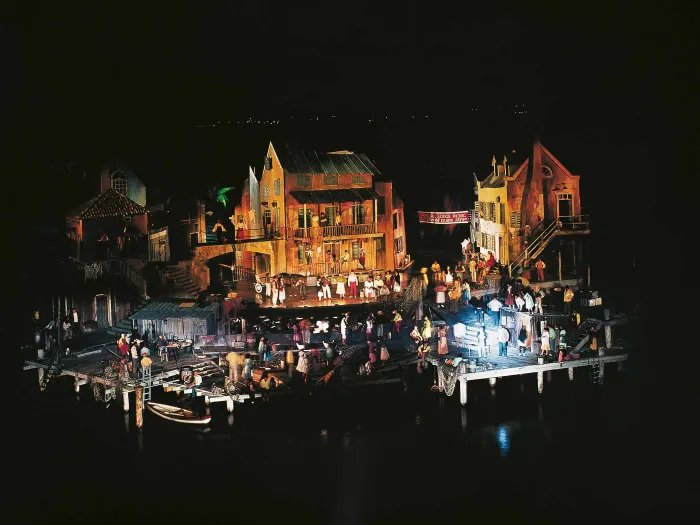 Szene aus "Porgy and Bess", 1971 auf der Seebühne, eine Plattform wird seitlich von zwei kleineren, tiefer liegenden Plattformen flankiert, die über Treppen zugänglich sind, im Zentrum steht ein dreistöckiges Haus, das in rötlichem Licht angestrahlt wird; rechts und links davon befindet sich jeweils ein weiteres Haus, zahlreiche Menschen sind auf der Bühne verteilt