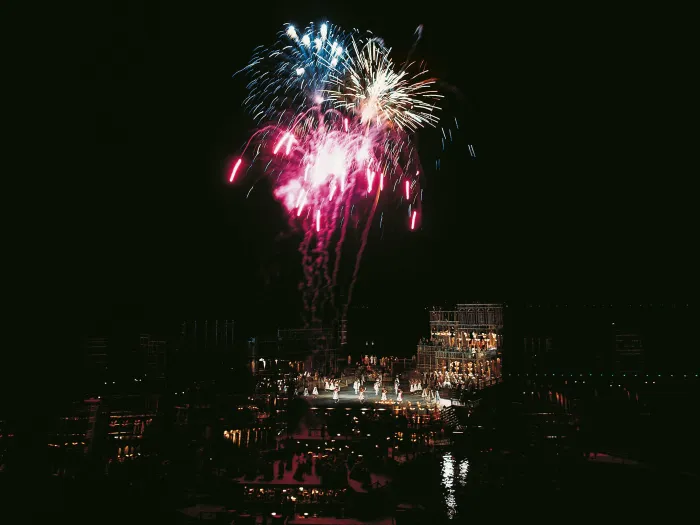 Szene aus "Eine Nacht in Venedig", 1975 auf der Seebühne, dreistöckiges Gebäude, viele Darstellende in Kostümen, am Himmel ist ein buntes Feuerwerk über dem Bühnenbild