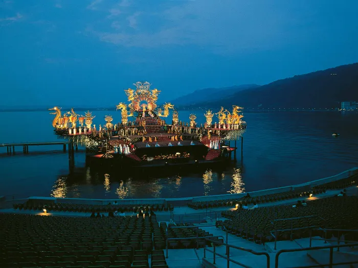 Szene aus "Turandot", 1979 auf der Seebühne, tribünenartiges Bühnenbild mit kostümierten Darstellenden in verschiedenen Ebenen, am Bühnenrand verlaufen goldfarbene Verzierungen mit Drachenfiguren