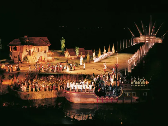 Szene aus "Der Zigeunerbaron", 1982 auf der Seebühne, auf der linken Seite befindet sich ein großes Haus, viele Darstellende in historischen Kostümen, ein Schiff im Wasser im Vordergrund, nahe an der Bühne, im Hintergrund beleuchtete Fontänen, die zu einer beleuchteten Gebäudesilhouette führen