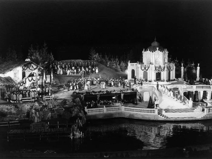 Szene aus "Der Vogelhändler", 1984 auf der Seebühne, zweistöckiges Bühnenbild, auf der oberen Ebene links befindet sich ein Gebäude und rechts ein zweistöckiger Pavillon, vor dem Pavillon führt eine Treppe in die untere Ebene, in der Mitte der Bühne sind viele kostümierte Darstellende 