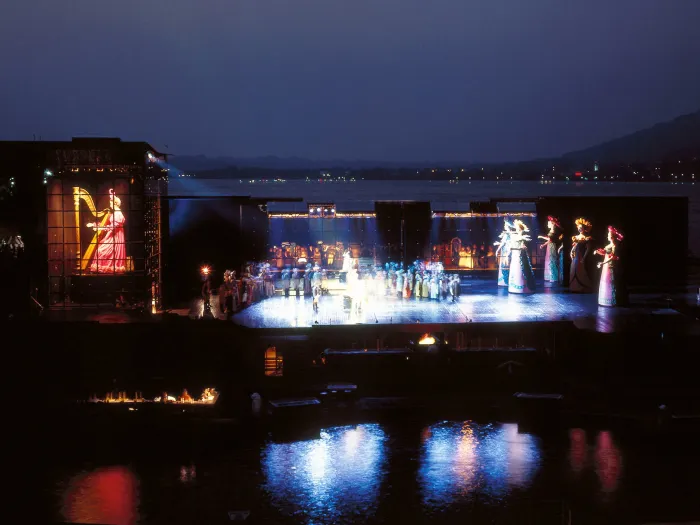 Szene aus "Hoffmanns Erzählungen", 1988 auf der Seebühne, rechts befinden sich mehrere übergroße Puppen in Blumenkostümen, links ist ein übergroßer Bildschirm mit einer Harfenspielerin, in der Mitte viele kostümierte Darstellende