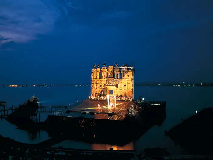 Szene aus "Nabucco", 1994 auf der Seebühne, große Gebäudefassade im Hintergrund, viele kostümierte Darstellende in einer reihe davor, auf der freien Fläche im Vordergrund befindet sich ein großes Reliefbild mit Abbild eines Menschen 