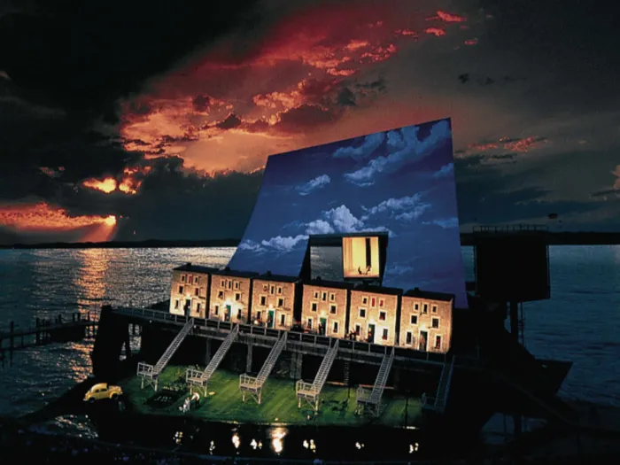 Szene aus "Fidelio", 1995 auf der Seebühne, sechs Häuserfassaden mit Treppen zu einer tieferliegenden Ebene, im Hintergrund großes Wolkenbild mit beleuchteter viereckiger Öffnung
