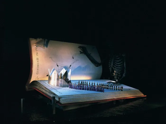 Szene aus "Ein Maskenball", 1999 auf der Seebühne, riesiges aufgeschlagenes Buch als Bühne mit einer großen silberfarbenen Krone links von der Bühne und einer großen Skelettfigur im Hintergrund, zwei Reihen kostümierter Darstellenden in Richtung der Krone 