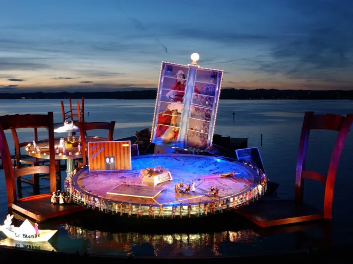 Szene aus "La Bohème", 2001 auf der Seebühne, großer Tisch als Bühnenfläche mit zwei überdimensionierten Stühlen im Wasser, im Hintergrund ein Kartenhalter mit Postkarten, die gemeinsam ein Gesamtbild ergeben und ein weiterer Tisch mit Stühlen, einige kostümierte Darstellende