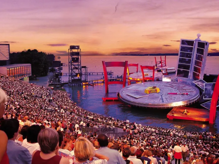 Szene aus "La Bohème", 2002 auf der Seebühne, großer Tisch als Bühnenfläche mit zwei überdimensionierten Stühlen im Wasser, im Hintergrund ein Kartenhalter mit Postkarten, die gemeinsam ein Gesamtbild ergeben und ein weiterer Tisch mit Stühlen