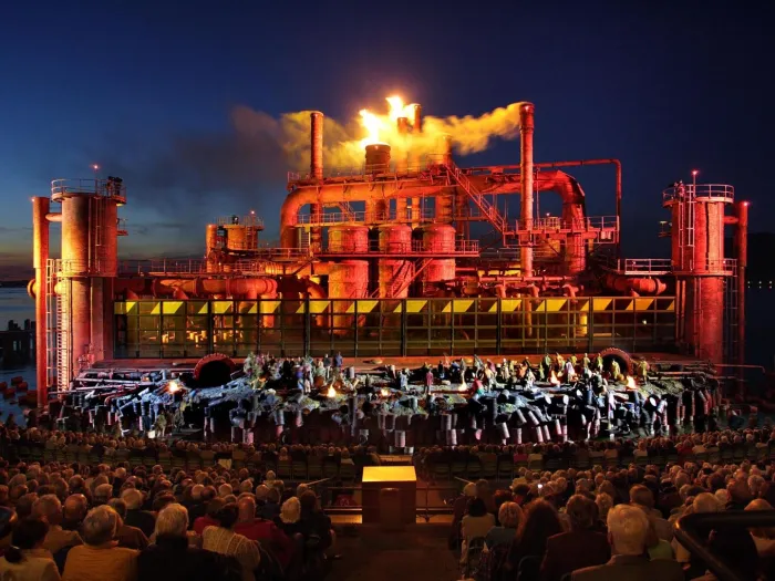 Szene aus "Der Troubadour", 2005 auf der Seebühne, Industrieanlage mit rauchenden Schornsteinen, einige kostümierte Darstellende im Vordergrund