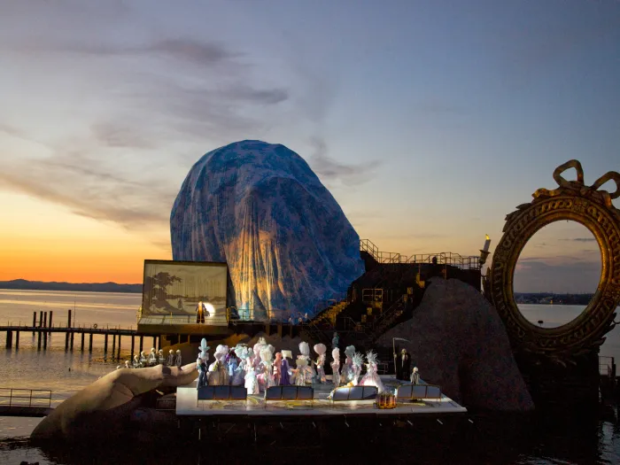 Szene aus "André Chénier", 2011 auf der Seebühne, große Hände umklammern die Seiten der Bühnenfläche, ein goldener Spiegelrahmen im Hintergrund und Figuren in historischen Kostümen auf der Bühnenfläche