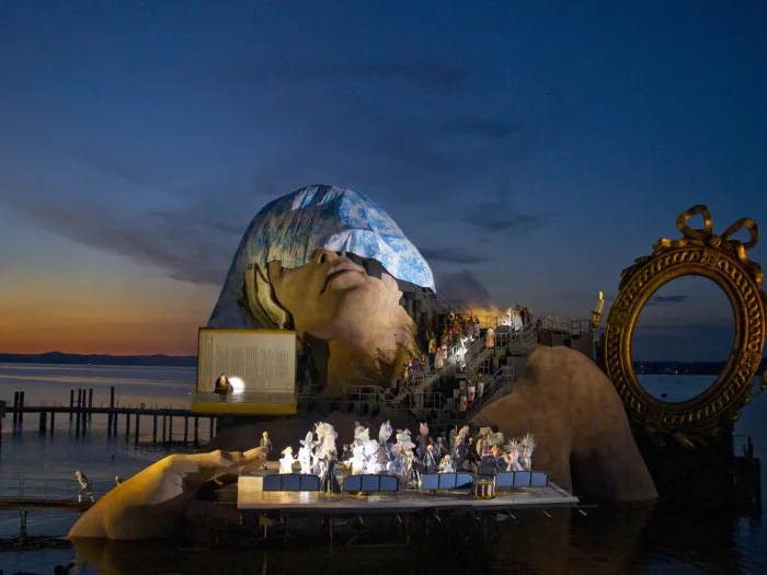 Szene aus "André Chénier", 2012 auf der Seebühne, große Hände umklammern die Seiten der Bühnenfläche, ein goldener Spiegelrahmen und einer riesiger mit einem Tuch bedeckter Kopf im Hintergrund und Figuren in historischen Kostümen auf der Bühnenfläche