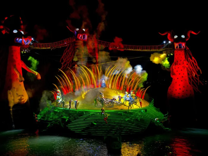 Szene aus "Die Zauberflöte", 2013 auf der Seebühne, im Hintergrund große rot beleuchtete Figuren mit Hörnern und durch Brücken verbunden, mittig eine grün-gelb beleuchtete runde Bühnenplattform, Rauch und mehrere kostümierte Darstellende im Vordergrund