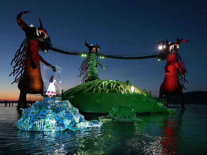 Szene aus "Die Zauberflöte", 2014 auf der Seebühne, drei große Drachenfiguren, durch Brücken verbunden, zwei Inseln im Wasser, im Vordergrund eine Person in einem transparenten Kasten