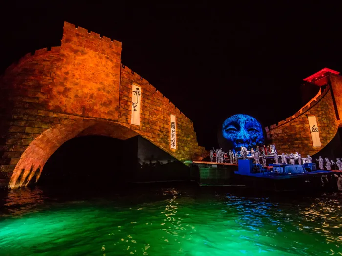 Szene aus "Turandot", 2015 auf der Seebühne, gebogener Steinbogen und großes blaues Gesicht im Hintergrund, im Vordergrund mehrere kostümierte Darstellende