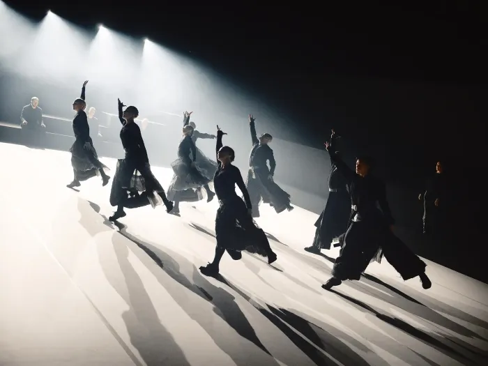 Sieben Tänzer in schwarzen Kostümen führen synchron eine Bewegung mit erhobenen Armen auf einer hell beleuchteten Bühne aus