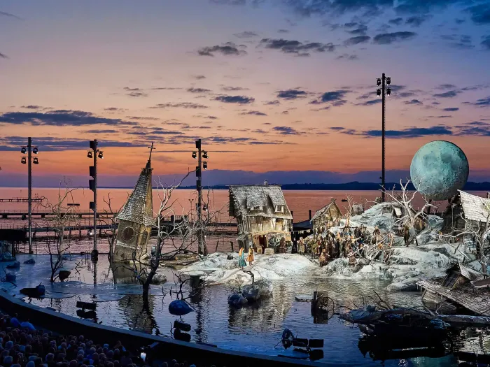 Kulisse "Der Freischütz" - Schneelandschaft überflutet, windschiefe Häuser, ein Kirchturm ragt aus dem Wasser, dramatischer Sonnenuntergang im Hintergrund in orange bis blau