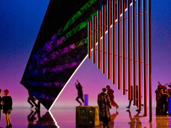 Szene aus "Achterbahn", 2011 im Festspielhaus, mehrere Darstellende tanzen, eine große Struktur hängt von der Decke, verspiegelter Boden, blau-pinker Hintergrund 