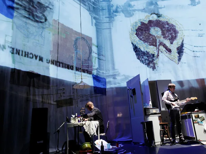 Szene aus "Home Work", 2011 im Festspielhaus, zwei Personen, eine am Tisch, die andere spielt Gitarre, Projektionen auf Leinwänden im Hintergrund, seitliche Aufnahme