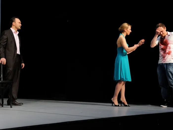 Szene aus "Waisen", 2011 im Theater Kosmos, drei kostümierte Darstellende, links ein Mann in schwarzem Anzug und weißem Hemd neben schwarzem Stuhl, rechts eine Frau in türkisem Kleid gestikuliert zu einem Mann mit kunstblutbeflecktem weißem T-Shirt