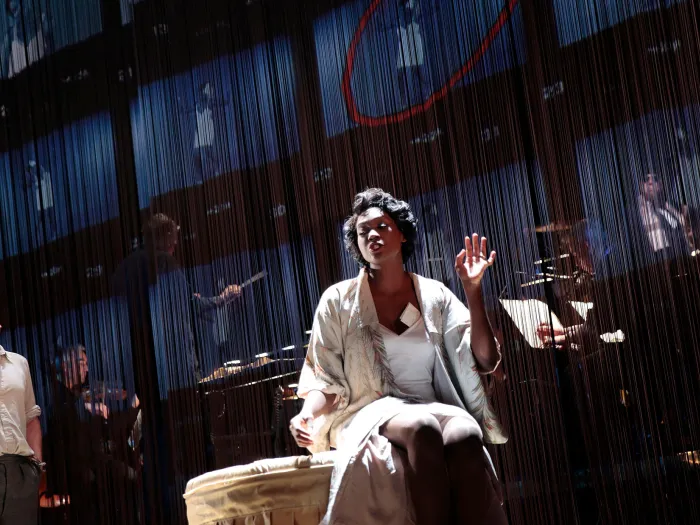 Szene aus "American Lulu", 2013 im Festspielhaus, zwei Darstellende, im Vordergrund eine Darstellerin sitzt und hebt eine Hand, links ein weiterer Darsteller, im Hintergrund von der Decke hängende Fäden und Musiker:innen